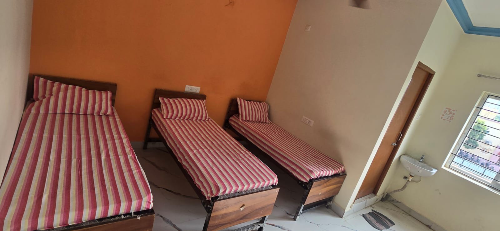 Triple sharing room with three beds, an orange accent wall, a washbasin and a window